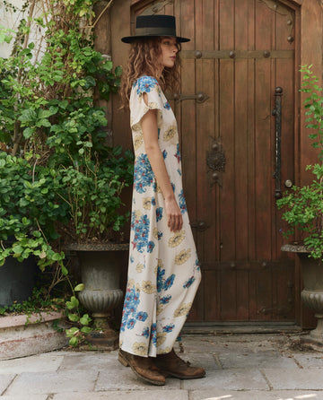 A woman in The Laurel Dress -- Sungold Floral by THE GREAT. PF25 stands sideways near a wooden door, surrounded by green plants, wearing a black wide-brim hat and brown boots.