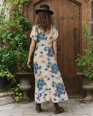 A person with curly hair wearing a black hat and THE GREAT. PF25 The Laurel Dress -- Sungold Floral stands facing a wooden door, surrounded by potted green plants.