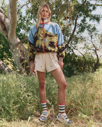 A person stands on grass wearing THE GREAT. SP26 TGO Embroidered Cargo Short in Cream with Black Daisies, a blue landscape-patterned jacket, striped socks, and sandals; green trees and sunlight are seen in the background.