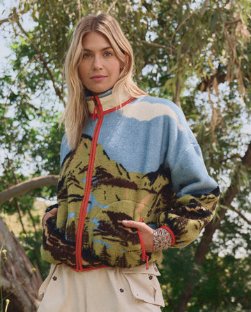 A woman stands outdoors before green foliage, wearing THE GREAT. SP26 TGO's Plush Terrain Full-Zip in California Eagle Landscape and light-colored pants.