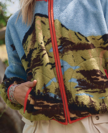 A person wearing THE GREAT. SP26 TGO The Plush Terrain Full-Zip – California Eagle Landscape, featuring a green, black, and blue landscape pattern with red trim, paired with beige pants. Only the lower face and upper body are visible.