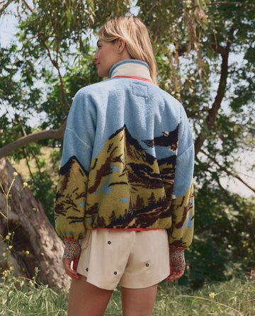 A person stands outdoors in THE GREAT. SP26 TGO "The Plush Terrain Full-Zip – California Eagle Landscape" jacket and cream shorts, surrounded by trees and plush terrain in the background.