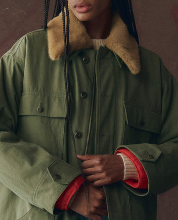 A person wears THE GREAT. F25 D1's The Quilted Liner Parka in Army with Hot Orange, featuring an oversized fit and removable faux fur collar, holding a section of their braided hair.