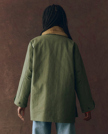 A person with long braids faces away, wearing THE GREAT. F25 D1 The Quilted Liner Parka in Army with Hot Orange and light blue jeans, set against a brown background.