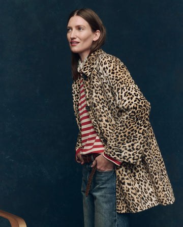 A woman wears THE GREAT. F25 COLLECTORS "The Car Coat." in Vintage Leopard over a red and white striped shirt and jeans, standing against a dark background.