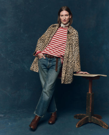 A woman stands with one hand on a small wooden table, wearing THE GREAT. SP25 COLLECTORS The Sailor Sweater in Maple Stripe, a leopard-print coat, blue jeans, and brown boots against a dark blue backdrop.