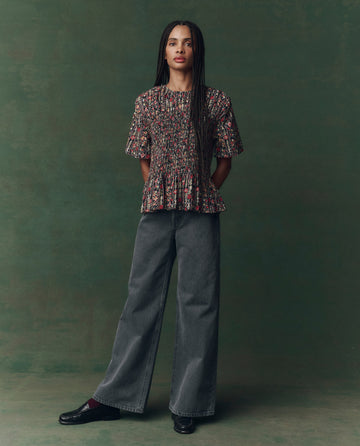 A person with long braids stands against a green backdrop, wearing THE GREAT.’s Smocked Flounce Top in Garland Print, paired with wide-leg gray jeans and black shoes.