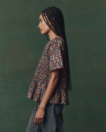 A person stands in profile against a green background, wearing THE GREAT. F25 D2's The Smocked Flounce Top – Garland Print and blue jeans.