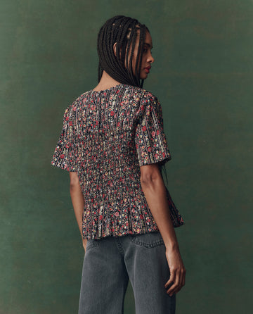 A woman with braids stands sideways, looking back, wearing THE GREAT. F25 D2 The Smocked Flounce Top in Garland Print and gray jeans against a green textured background.