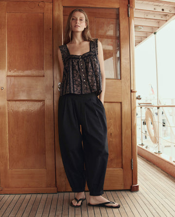 A woman stands on a wooden deck in front of double doors, wearing THE GREAT. SU26 The Victorian Pleat Tank in Black And Cream Token Floral, paired with loose dark pants and sandals.
