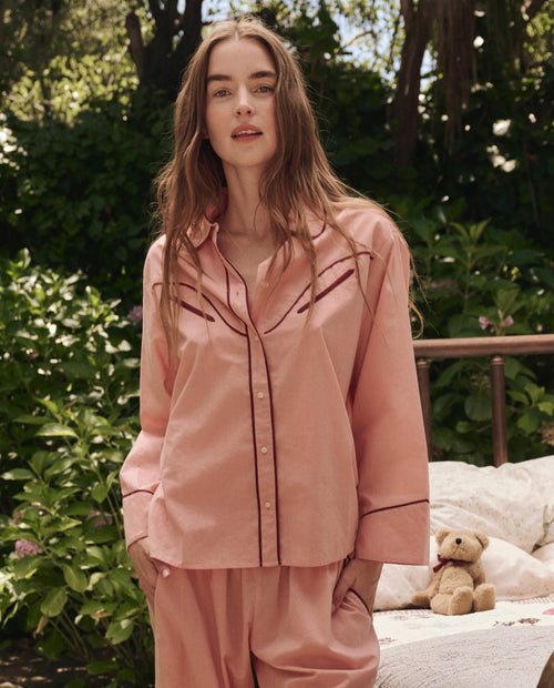 A woman wearing THE GREAT. SP25 SLEEP's The Western Pajama Shirt in Rose Bloom stands outdoors before a bed with green foliage and a teddy bear in the background.