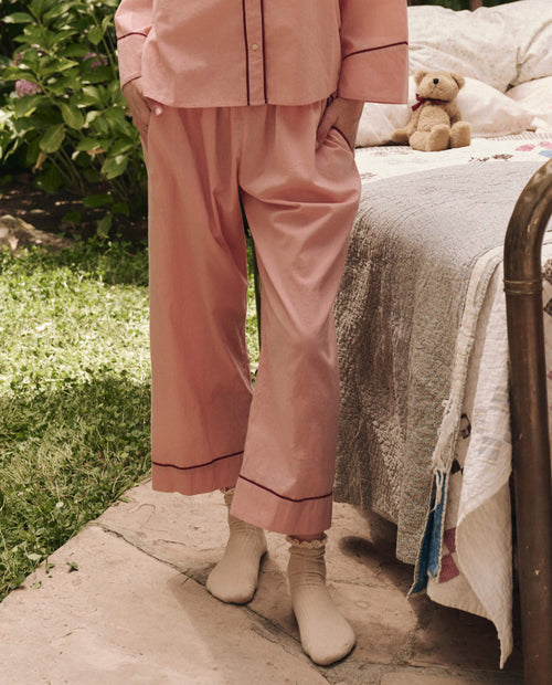 Wearing THE GREAT. SP25 SLEEP Western Easy Sleep Pant in Rose Bloom, a person in pink pajamas and cream socks stands by a bed with a quilt and teddy bear in an outdoor garden, enjoying effortless comfort.