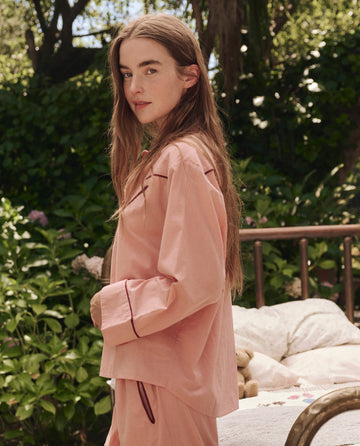 A person with long brown hair wears THE GREAT. SP25 SLEEP's The Western Pajama Shirt in Rose Bloom, standing outdoors by a bed with green foliage and trees in the background.