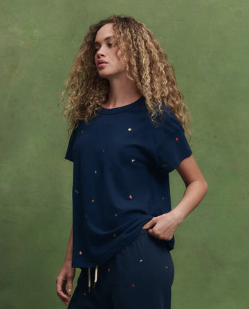 A woman with curly hair wears THE GREAT. SU26's The Boxy Crew W/ Tossed Floral Emb. in navy, paired with matching pants, as she stands against a green background.