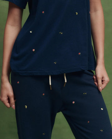 A person stands against a green background wearing THE GREAT. SU26 The Boxy Crew w/ Tossed Floral Embroidery in navy—boxy fit, small embroidered florals, and white drawstrings for a handcrafted Los Angeles vibe.
