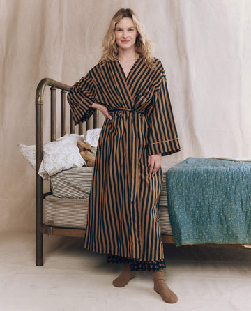 A person in THE GREAT. F25 SLEEP The Robe. -- Club Stripe stands by a vintage-style bed with patterned bedding and a teddy bear on the pillows.