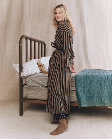 A woman in THE GREAT. F25 SLEEP The Robe. -- Club Stripe and socks stands beside a made bed with floral bedding, a blue blanket, and a stuffed animal.