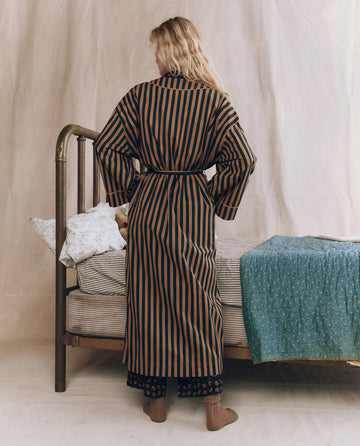 A person wearing THE GREAT. F25 SLEEP The Robe. -- Club Stripe stands facing away next to a vintage metal bed with a blue patterned blanket and floral pillowcases.