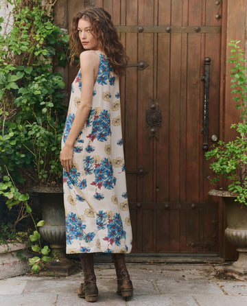 In front of a wooden door, a woman with curly hair models THE GREAT. PF25's Windward Dress in Sungold Floral—sleeveless summer fashion paired with brown boots, surrounded by lush greenery.
