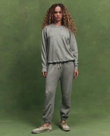 A woman with curly hair stands against a green background, wearing THE GREAT. SU26's The Stadium Sweatpant W/ Tossed Floral Emb. in Varsity Grey, paired with a matching sweatshirt and beige sneakers.
