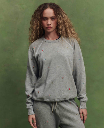 A person with curly hair wears THE GREAT.’s The College Sweatshirt w/ Tossed Floral Embroidery in Varsity Grey, standing in front of a green background.