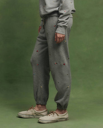 A person stands against a green backdrop wearing THE GREAT. SU26 The Stadium Sweatpant W/ Tossed Floral Emb. in Varsity Grey, paired with a matching sweatshirt and white sneakers.