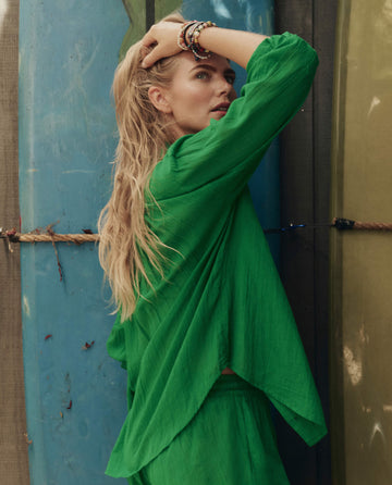 A woman with long blonde hair wears THE GREAT. SP26 SWIM The Cove Shirt in Bright Sprig, standing before surfboards and looking over her shoulder with one hand touching her head.