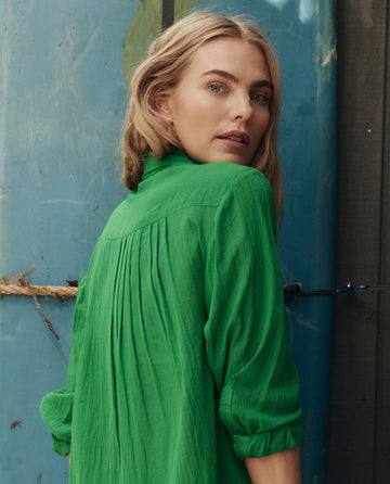 A woman with blonde hair wears THE GREAT. SP26 SWIM's The Cove Shirt in Bright Sprig, standing in front of a blue textured wall and looking over her shoulder at the camera.
