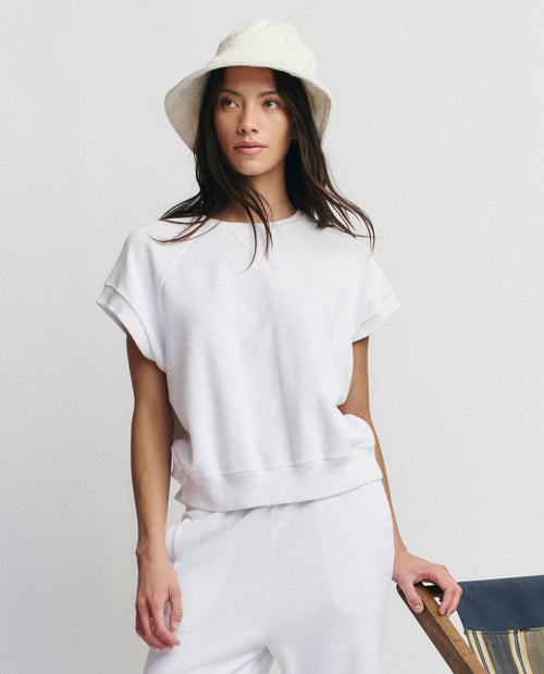 A woman wears THE GREAT. SU25 MICRO TERRY Wedge Sweatshirt in True White, posing next to a striped chair in a minimal setting.
