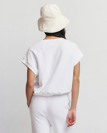 A person stands with their back to the camera, wearing THE GREAT. SU25 MICRO TERRY The Micro Terry Wedge Sweatshirt in True White, paired with white pants and a white bucket hat against a plain background.