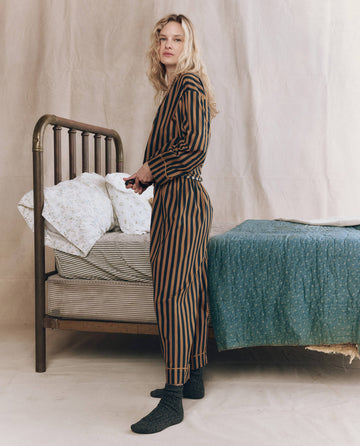 Wearing THE GREAT. F25 SLEEP's The Easy Sleep Pant in Club Stripe, a person stands by an unmade bed with patterned bedding and a green blanket in a softly lit, neutrally decorated room.