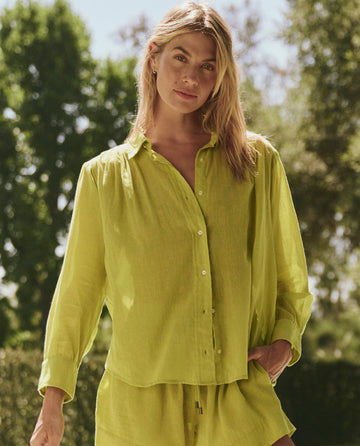 A woman stands outdoors among greenery, wearing THE GREAT. SP25 SWIM The Cove Shirt in Citron with matching shorts.