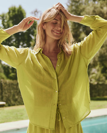 A person wearing THE GREAT. SP25 SWIM's The Cove Shirt in Citron with matching pants stands poolside outdoors, smiling with eyes closed and hands raised to their head, surrounded by trees and greenery.