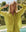 A person wearing THE GREAT. SP25 SWIM's The Cove Shirt in Citron with matching pants stands poolside outdoors, smiling with eyes closed and hands raised to their head, surrounded by trees and greenery.