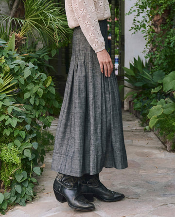 Person stands on a stone path among greenery, wearing THE GREAT. PF25 The Rhythm Skirt – Smoke Wash, black boots, and a beige long-sleeve top—a timeless look for understated style.