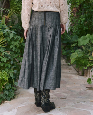 A person stands on a stone path in a light knit top and THE GREAT. PF25 Rhythm Skirt—Smoke Wash, a long gray pleated skirt with back zipper—plus black patterned boots, surrounded by lush greenery. Fashion meets nature in this serene scene.