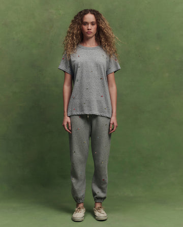 A woman faces forward against a green background, wearing THE GREAT. SU26's The Boxy Crew W/ Tossed Floral Emb. in Heather Grey with matching sweatpants and beige sneakers.