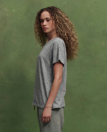A person with curly hair stands in profile wearing THE GREAT. SU26 The Boxy Crew W/ Tossed Floral Emb. in Heather Grey against a green background.
