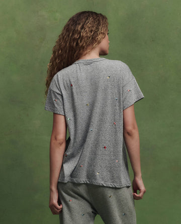 A person with long curly hair faces away, wearing THE GREAT. SU26's Boxy Crew W/ Tossed Floral Emb. in Heather Grey and pants with small colorful embroidered dots, set against a plain green background.