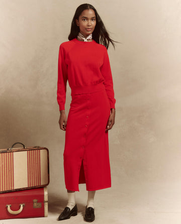 A woman in a red dress and patterned scarf stands beside vintage suitcases, wearing black shoes and carrying "The Crew Neck Sweater" by THE GREAT. SP25 HIGH TWIST YARN draped over her arm.