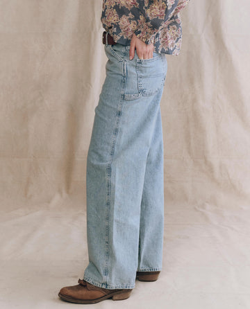 A person stands on a light surface wearing The Mill Jean – Dakota Wash by THE GREAT. PF25, brown shoes, and a floral long-sleeve shirt, with one hand in their back pocket.