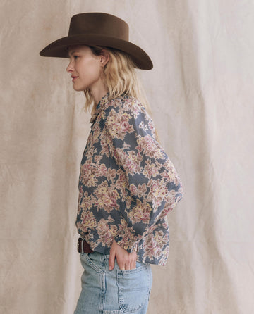 A person in a brown wide-brimmed hat and light blue jeans wears THE GREAT. PF25's The Hemingway Top in Indigo Floral, standing in profile against a beige fabric backdrop.