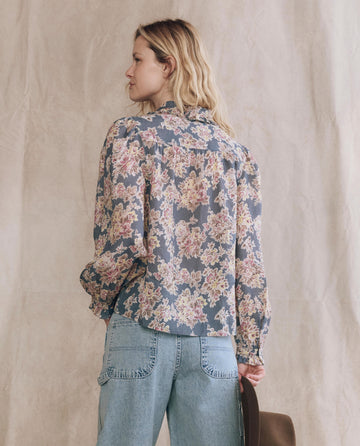 A person faces away, wearing THE GREAT. PF25 The Hemingway Top in Indigo Floral with light blue jeans, holding a brown hat in their left hand against a neutral background.