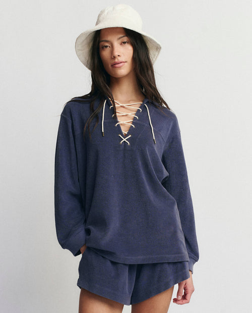 A person wears THE GREAT. SU25 MICRO TERRY’s The Micro Terry Lace Up Tunic in Vintage Blue with matching shorts and a white bucket hat, standing against a plain light background.
