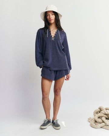 A woman stands indoors next to a coiled rope, wearing a white hat, sneakers, and THE GREAT. SU25 MICRO TERRY Micro Terry Swing Short in Vintage Blue with a matching navy lace-up long-sleeve top.