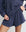Wearing THE GREAT. SU25 MICRO TERRY’s Micro Terry Swing Short in Vintage Blue, a person stands with one hand on their hip and the other adjusting the waistband, pairing it with a loose-fitting, long-sleeve dark blue top.