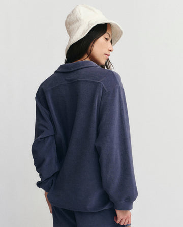 A person wearing THE GREAT. SU25 MICRO TERRY The Micro Terry Lace Up Tunic in Vintage Blue stands facing away, looking to the side against a plain light background.