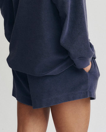 A person wears THE GREAT. SU25 MICRO TERRY The Micro Terry Swing Short in Vintage Blue, paired with a loose, long-sleeve navy top and has one hand in their pocket, shown from mid-thigh to waist.