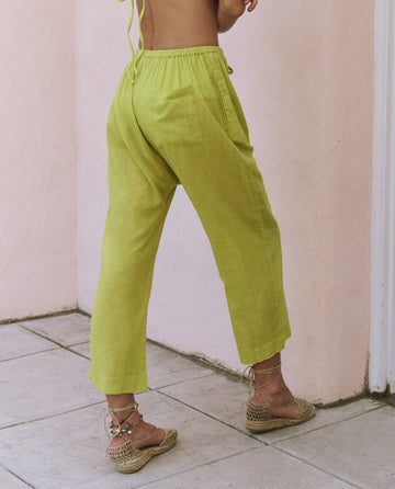 A person wearing THE GREAT. SP25 SWIM The Reef Pant in Citron and woven sandals stands on a tiled floor near a light pink wall, showcasing stylish summer clothing.