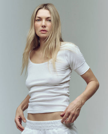 A woman with long blonde hair wears THE GREAT. SU25 BABY RIB's The Baby Rib Ballet Tee in True White and white pants, standing against a plain light background while looking at the camera.